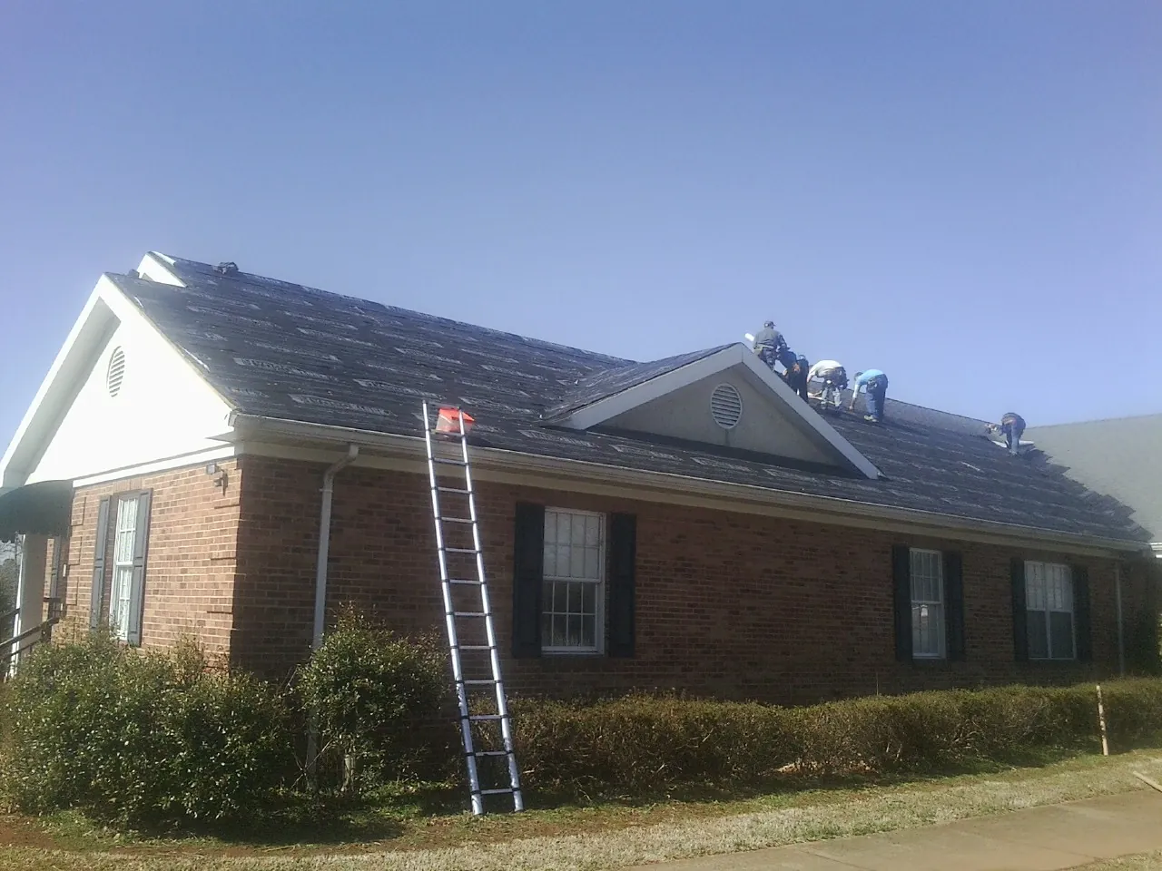 Crew tearing shingles off a brick ranch roof with a ladder set — Emergency Roof Repair work by our crew in Grace