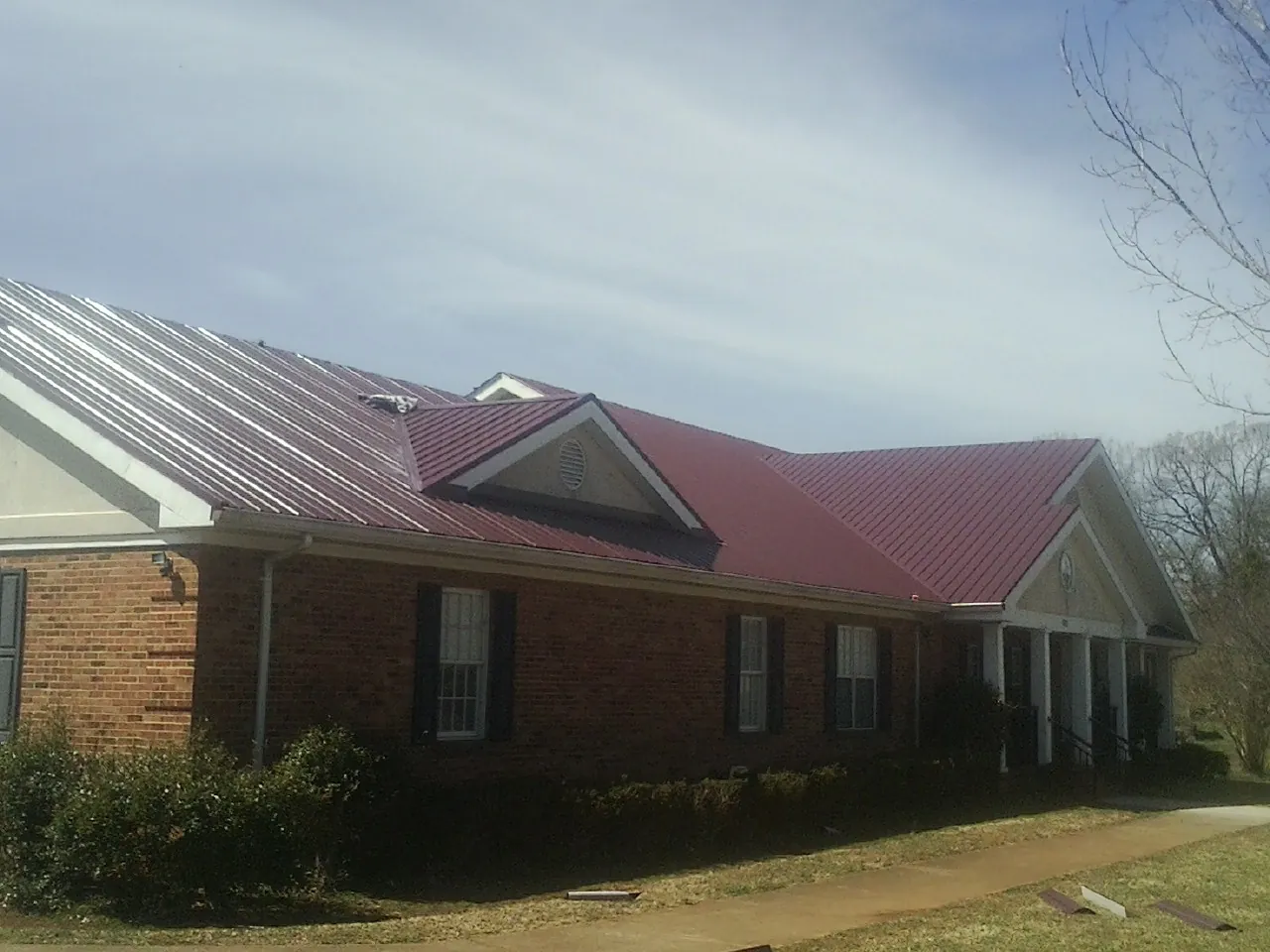 Red metal roof with gabled entries on a long brick building, from a Hail Damage Roof Repair job in Grace