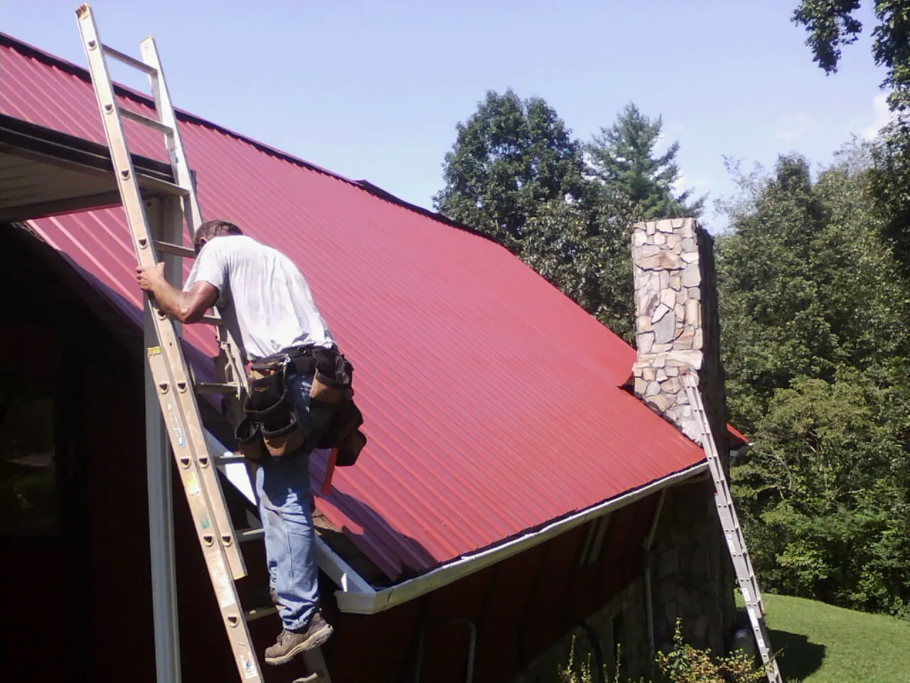 Roofer on a ladder beside a red metal roof and stone chimney — Soffit & Fascia Repair by Roofing Brothers in Grace
