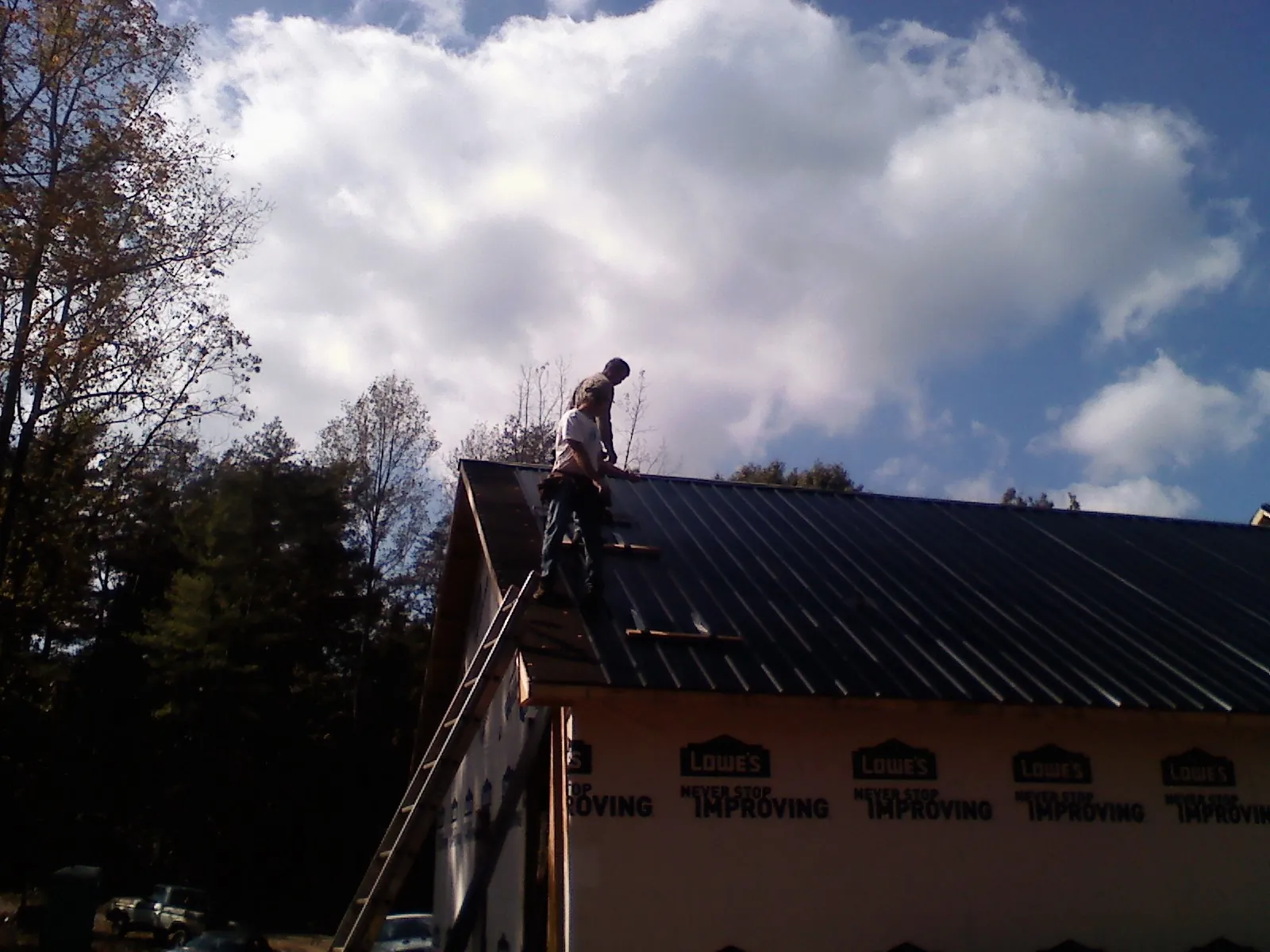 Roofer fastening dark metal panels high against a cloudy sky, from a Roof Inspection job in Grace